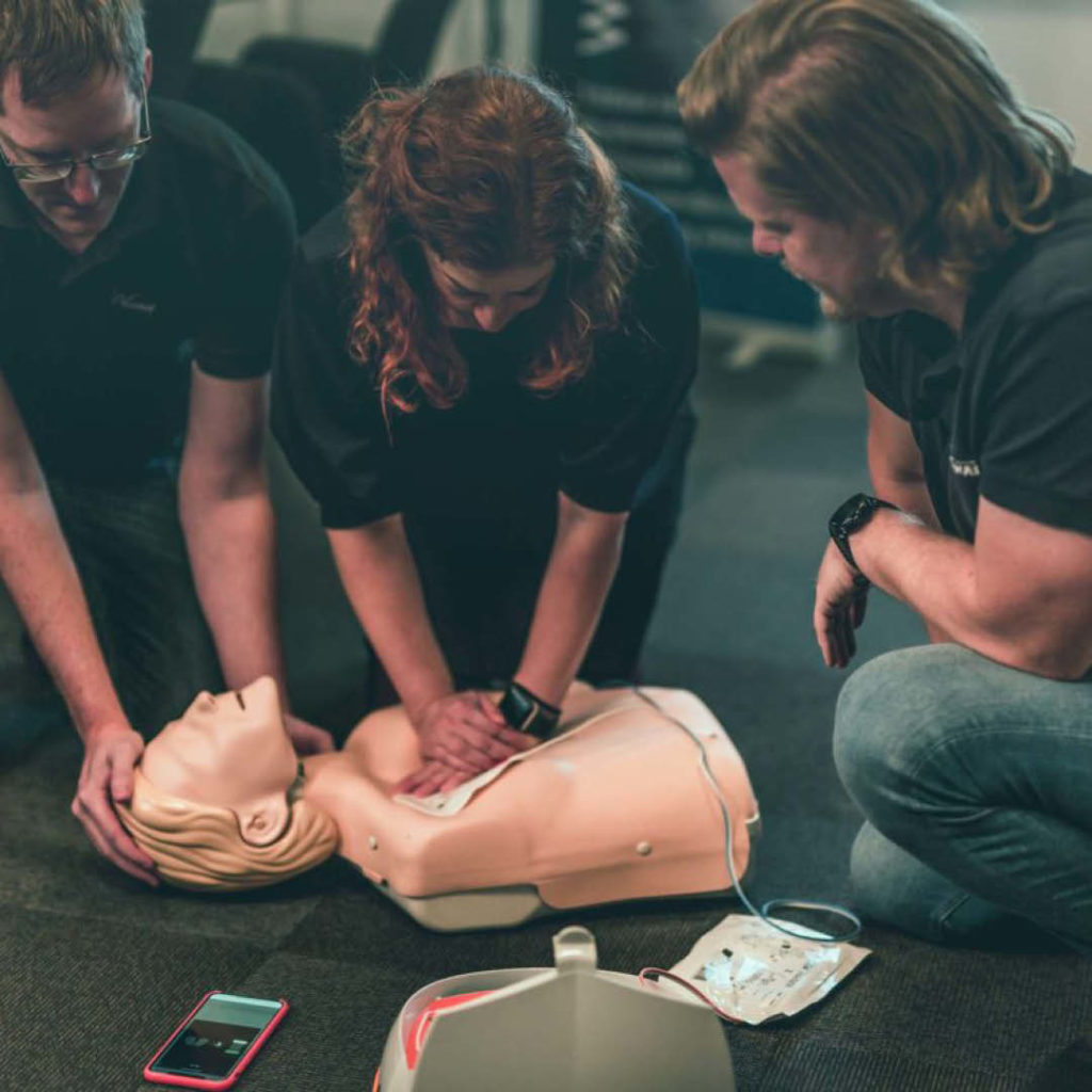 En Anna-dukke ligger på gulvet, en kursdeltager utfører HLR og en hjertestarter er påkoblet. En Blostrupmoen-instruktør observerer