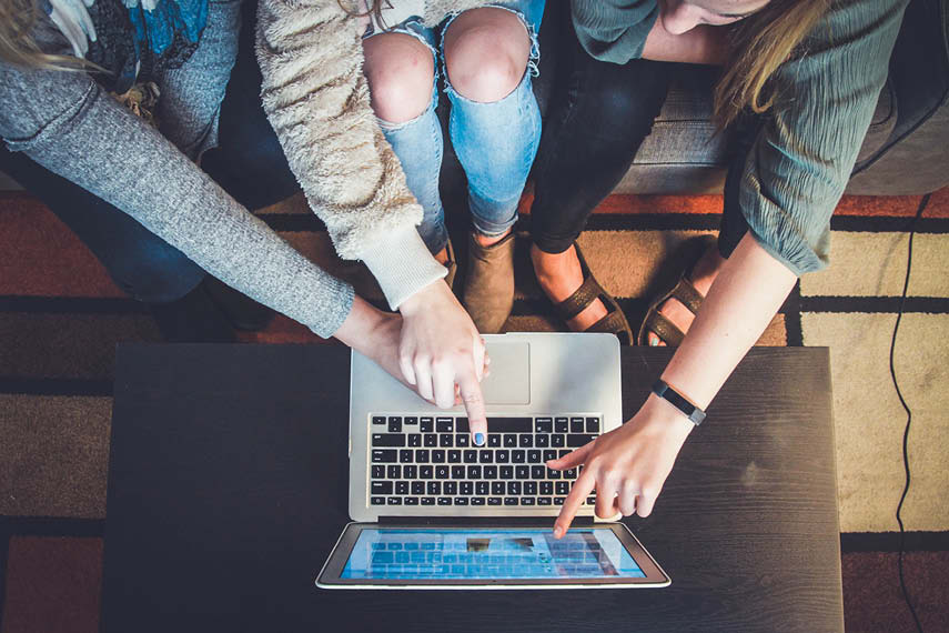 Tre mennesker peker på skjermen på en PC. Bildet er tatt ovenifra og viser kun bena og armene på menneskene.