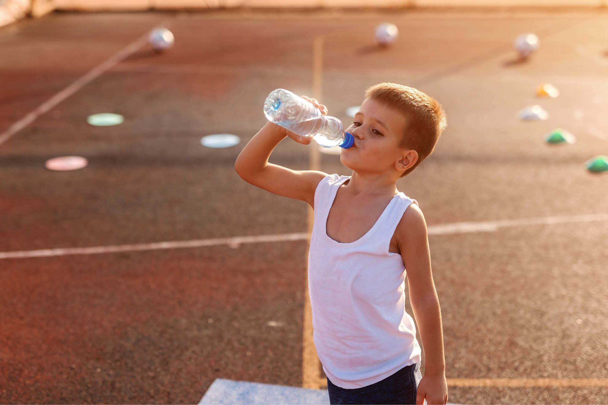 en liten gutt står å drikker av en vannflaske på en løpebane etter trening. Han har på seg singlet som indkerer at det er en varm dag.