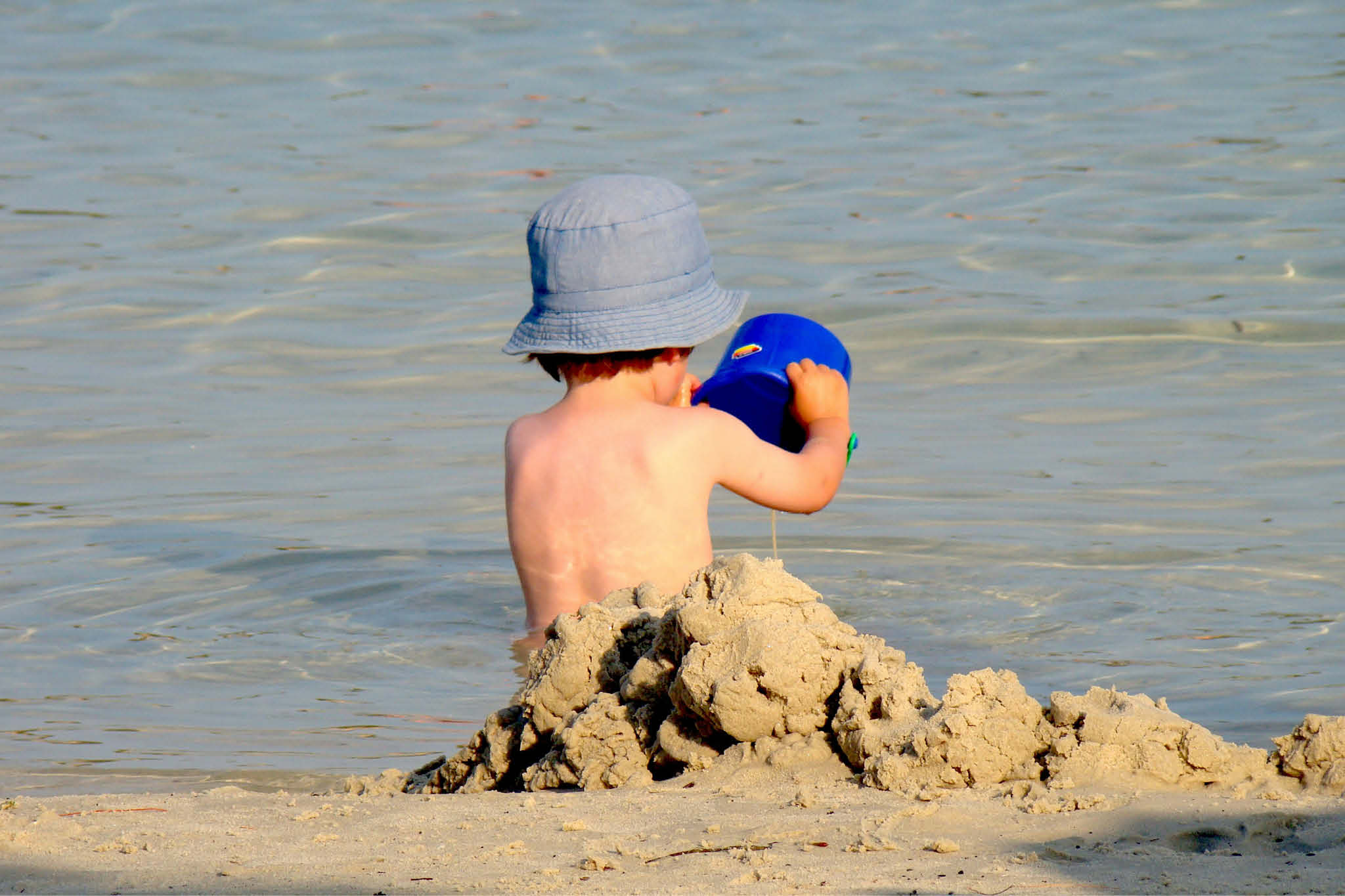 Et barn sitter på stranden iført solhatt og bygger sitt første sandslott.