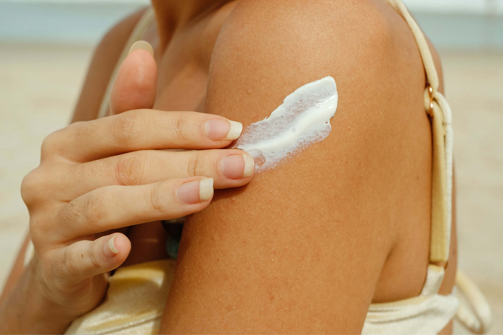 En kvinne smører solkrem på armen sin, hun er på stranden i strålende solskinn