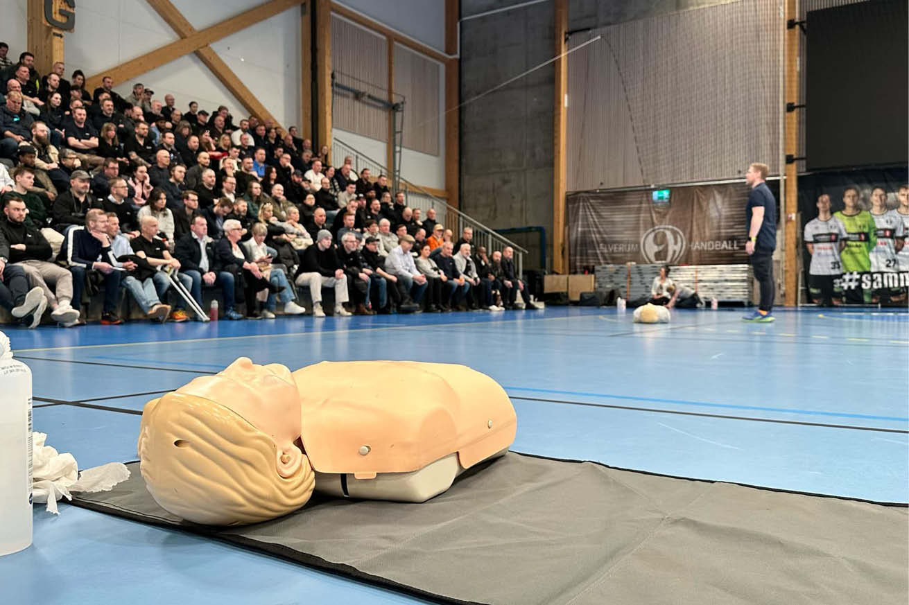 Mange mennesker sitter på tibunen og hører på en instruktør fra Blostrupmoen undervise i førstehjelp. i bilde ser du også en treningsdukke